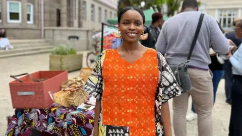 BBC Seun smiles at the camera next to a stall with a traditional African cover over it with baskets on top of it