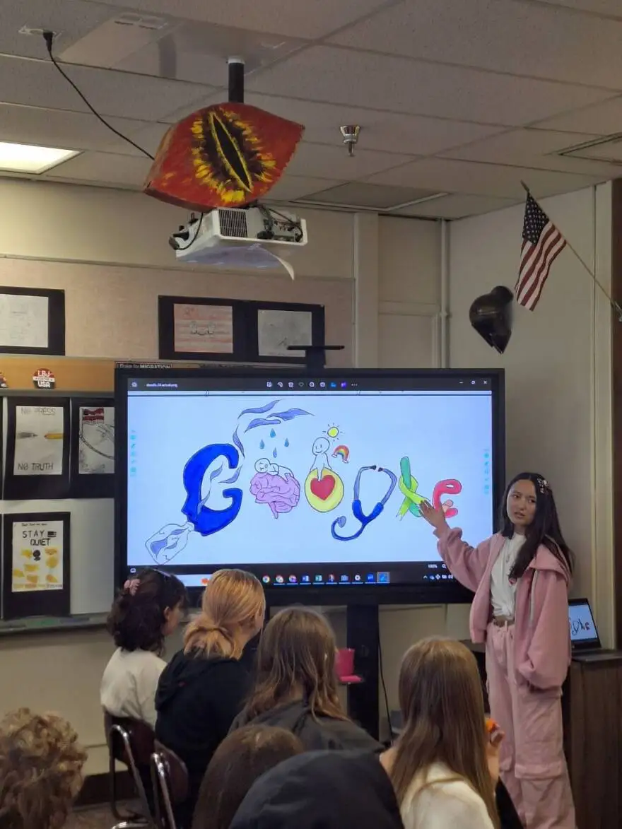 A middle school girl wearing a pink sweatshirt, white shirt and pink sweatpants stands in front of a big screen projecter with her art design on it. She is pointing to the design and talking to a classroom of other students.
