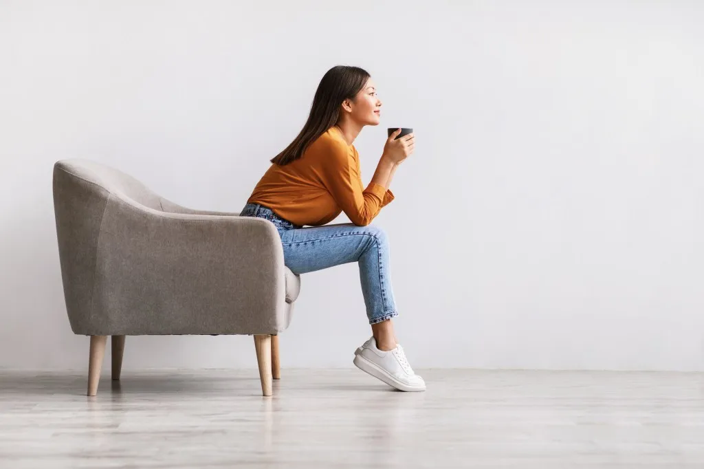 Young Asian woman sitting in armchair drinking hot coffee against a white wall