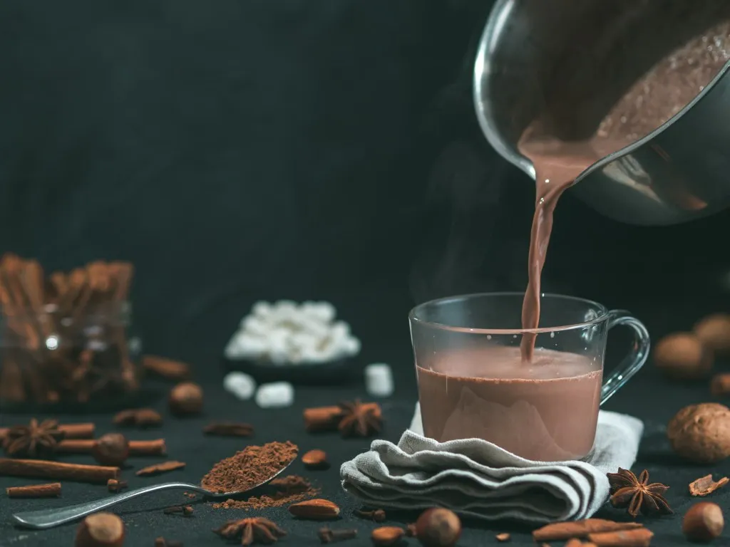 Pouring hot chocolate cocoa into a glass mug on a black table with ingredients around