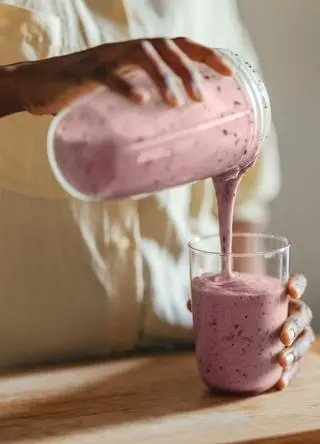 Close-up of hands of a woman wearing beige shirt and pouring smoothie into a glass on a kitchen table.