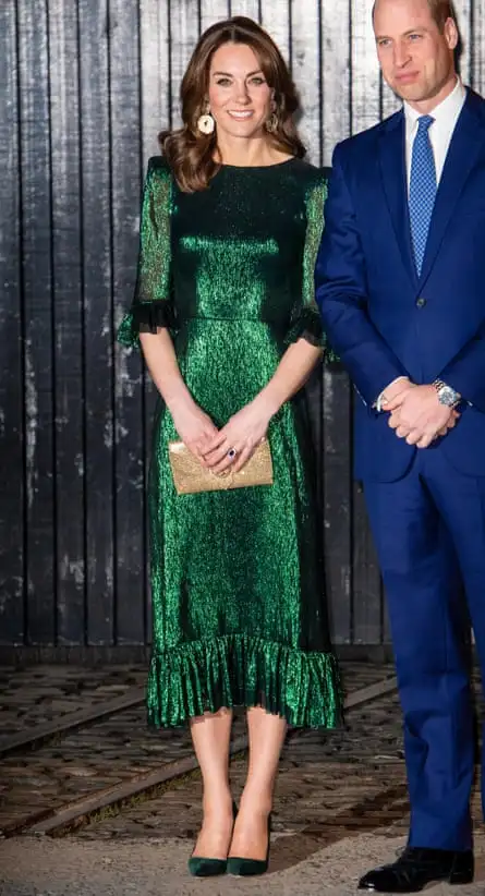 Kate seen full-length in a long, dark green glittery version of the dress; it has ruffles at the elbows and hemline, and she is wearing matching high-heeled shoes and clutching a small gold handbag. Her long dark hair is loose and waved, and she is wearing large gold earrings. Prince William is partly seen next to her, wearing a dark blue suit, white shirt and tie; they are standing against what appears to be a wall or partition of thin, vertical black planks of wood. 