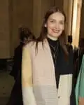 Roksanda Ilinčić, photographed at London fashion week, February 2022: she is wearing a black high-necked top with three gold necklaces, the lowest one a cross, under a collarless oversized jacket with panels in shades of pale gold, cream and grey with narrow stripes and a shoulder in black; it is fastened with a single button at waist level. She has straight, brown hair worn loose and is wearing bright red lipstick and smiling at the camera. 