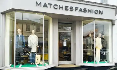 A double-fronted shop with mannequins in the window and signage promoting the Raey brand; the shop is painted in pale grey and the door is shut. The mannequins are wearing dark blue, grey, khaki and cream garments including coats, jeans, jackets and trousers