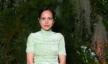 Anna Jewsbury at the British Vogue x Netflix Bafta party in London, May 2024: she has dark brown hair tied back to show the multiple pairs of ornate silver, gold and pearl earrings she is wearing; she wears a pale silvery-green high-necked and short-sleeved velvet top with a leaf-like pattern and is looking straight at the camera. She has dark brown eyes and is wearing pink lip gloss; she stands against a dark backdrop of trailing plants and leaves