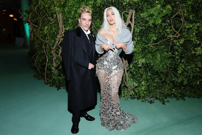 The designer John Galliano in his 60s in a black suit with Kim Kardashian in a silver dress and cardigan at the 2024 met gala