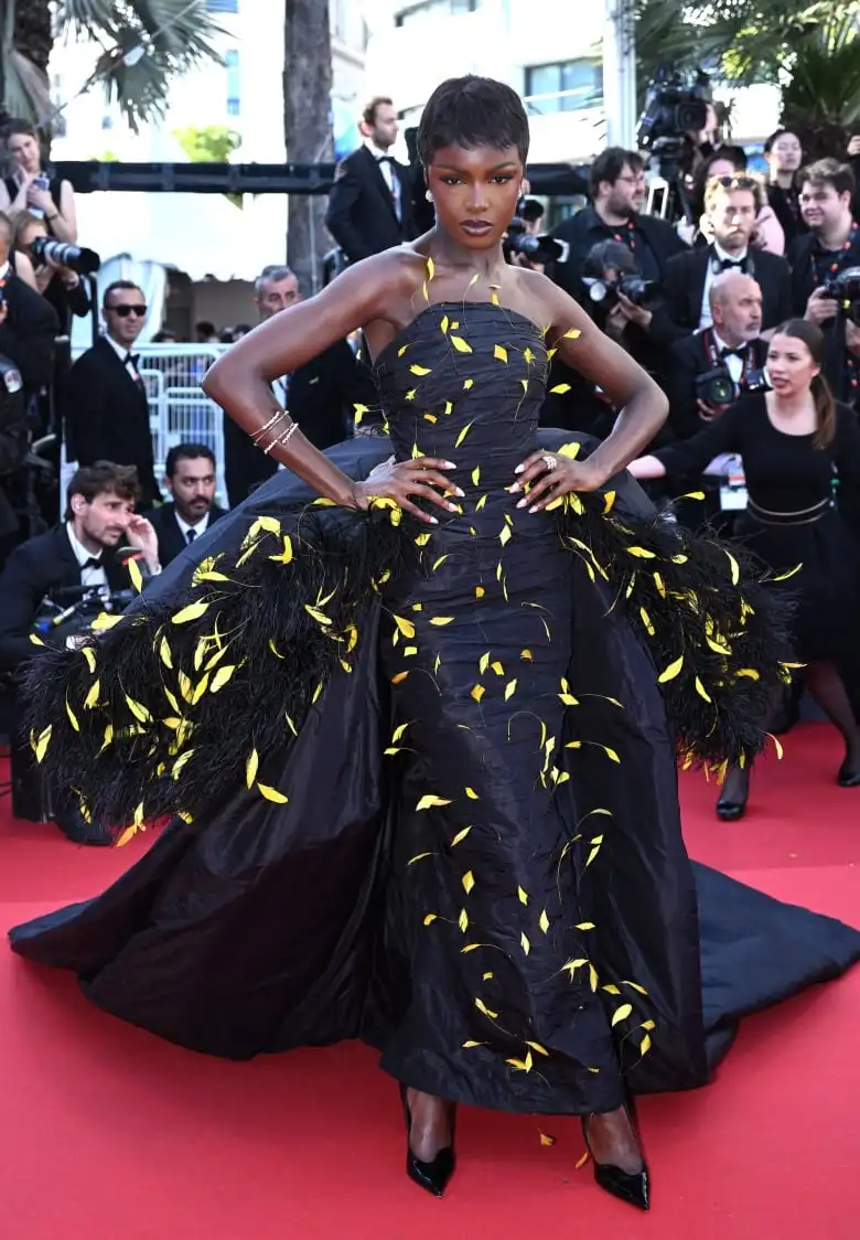 Leomie Anderson on the red carpet wearing a black, strapless taffeta gown adorned with black and yellow feathers.