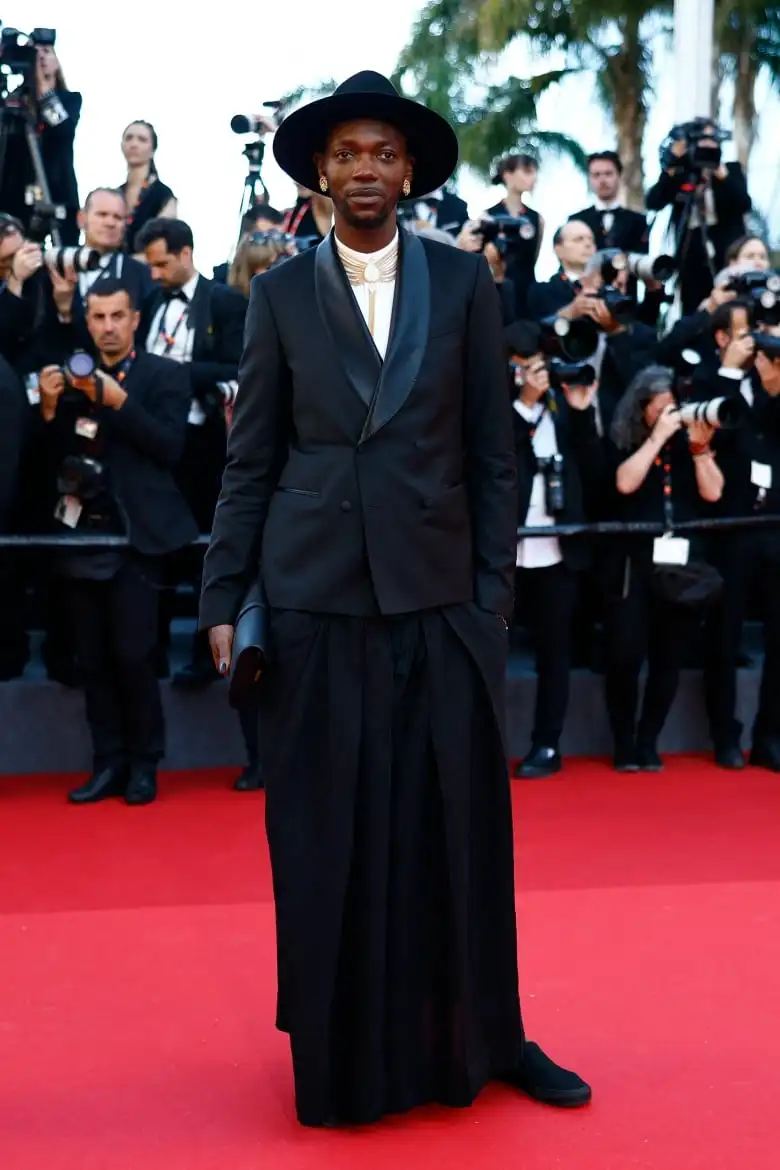 Baloji on the red carpet wearing a black skirt suit with a black tuxedo jacket and hat.