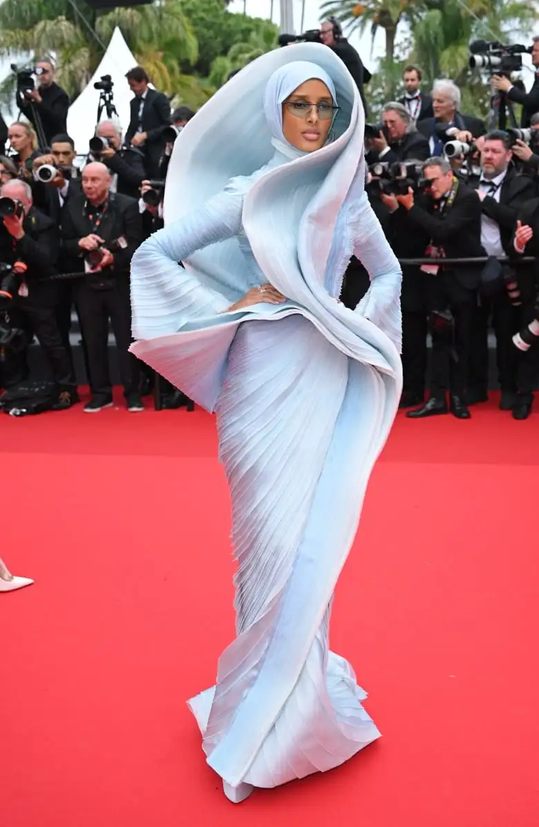 Rawdah Mohamed on the red carpet wearing a light blue gown that has a sculptural piece that curves up her body and flows around her head. 