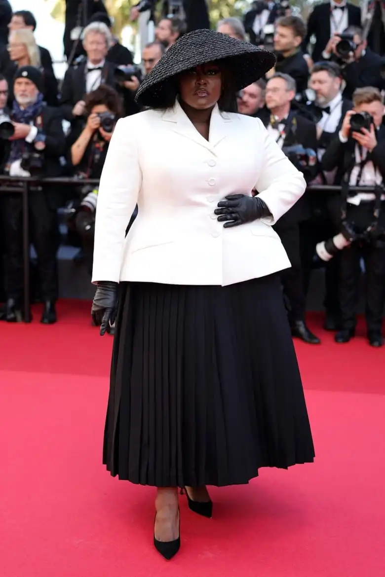 Yseult on the red carpet wearing a skirt suit with a white jacket, black skirt, black leather gloves, pumps and hat.