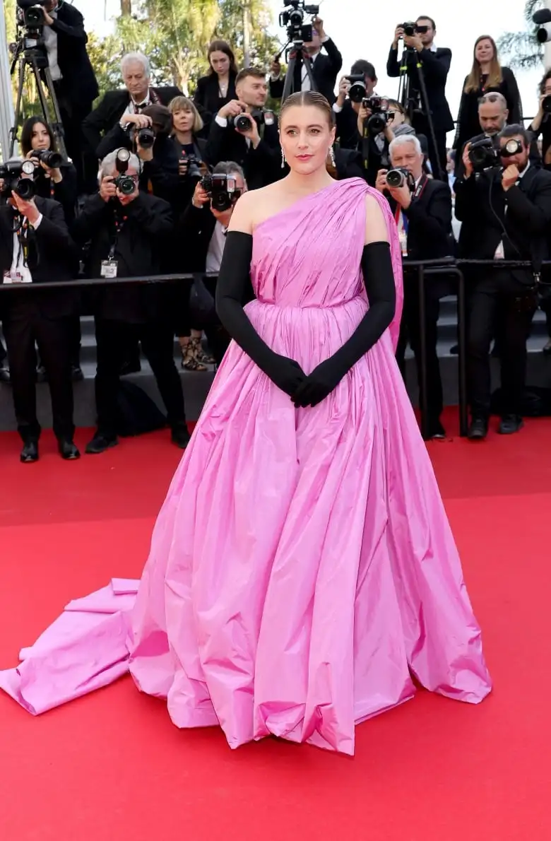 Greta Gerwig on the red carpet wearing a bright pink gown and black opera gloves.