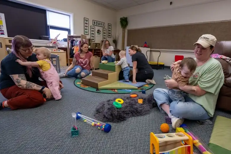 Several people are seen sitting on the floor of a playroom with babies.