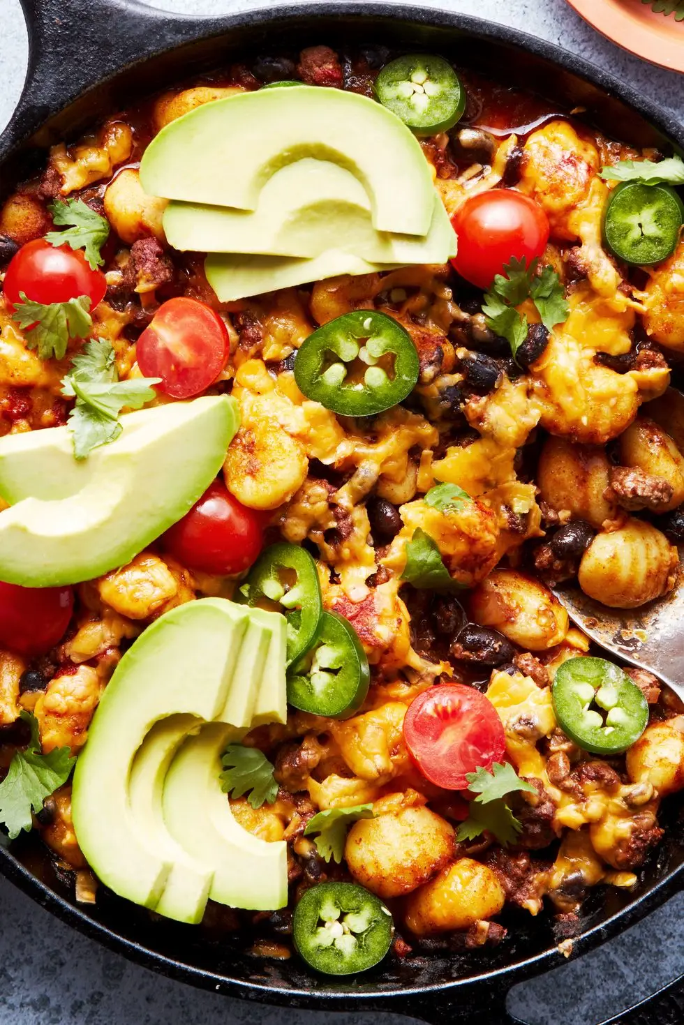 a pan with gnocchi covered in enchilada sauce, cheese, avocado, cilantro, and cherry tomatoes