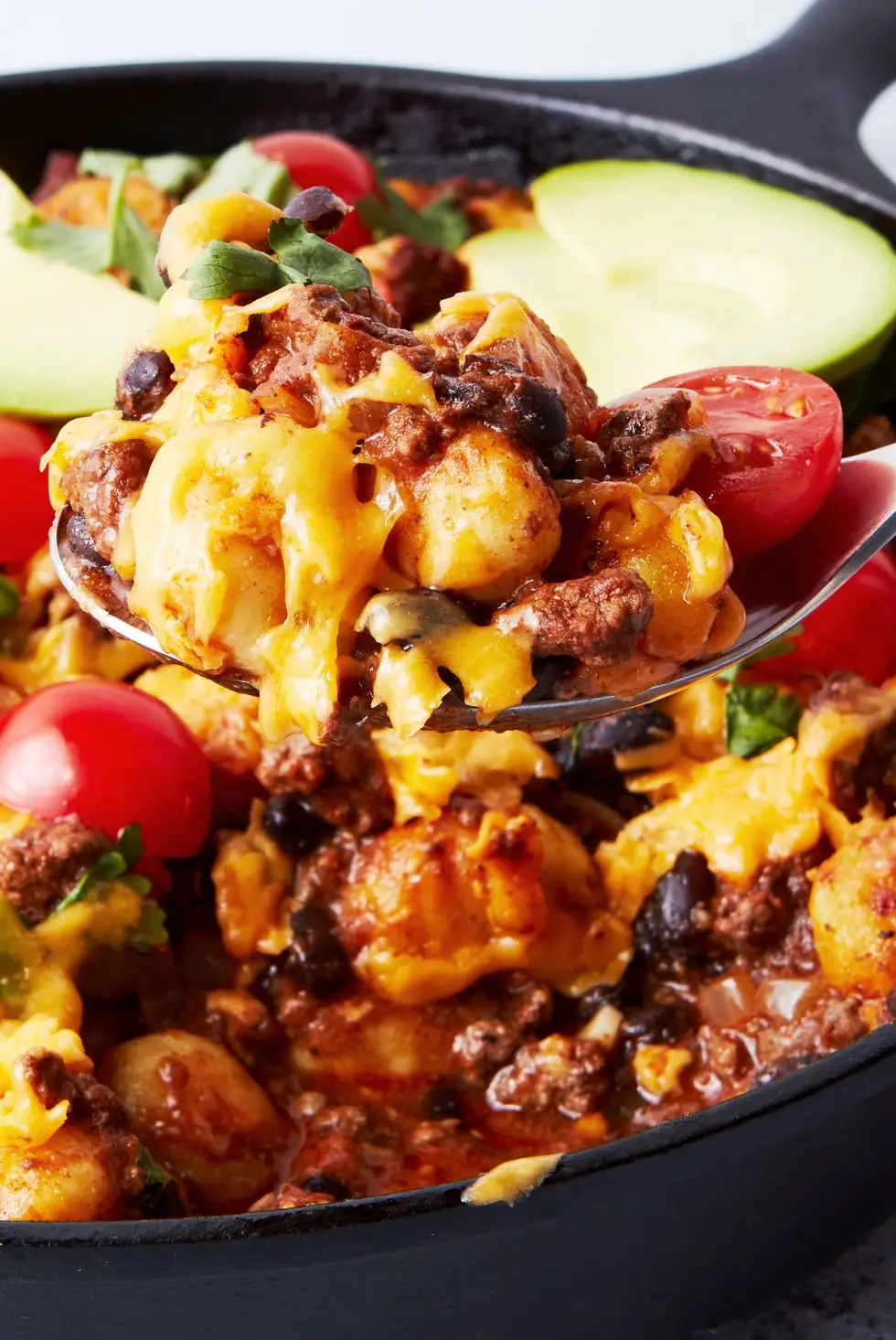 a pan with gnocchi covered in enchilada sauce, cheese, avocado, cilantro, and cherry tomatoes