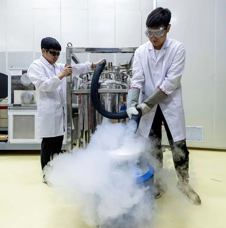 Two people wearing white laboratory coats, gloves, and goggles, standing on either side of large vat pouring liquid nitrogren