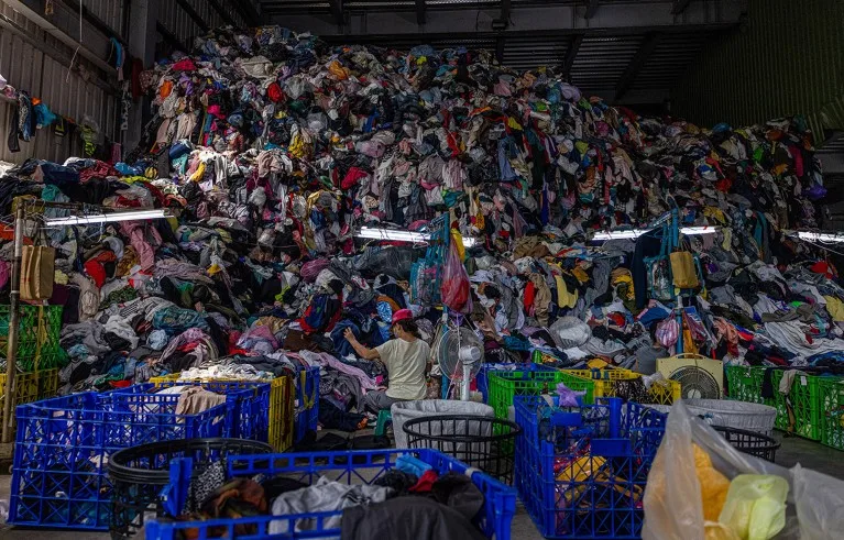 A single worker stands beneath a vast pile of unsorted second-hand clothes.