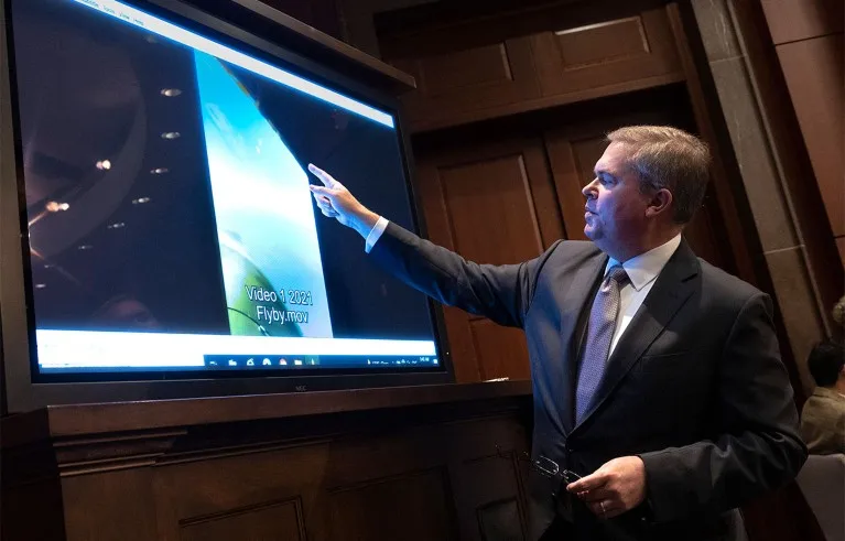 A man in jacket and tie points to a monitor playing a video of an unidentified aerial phenomena.