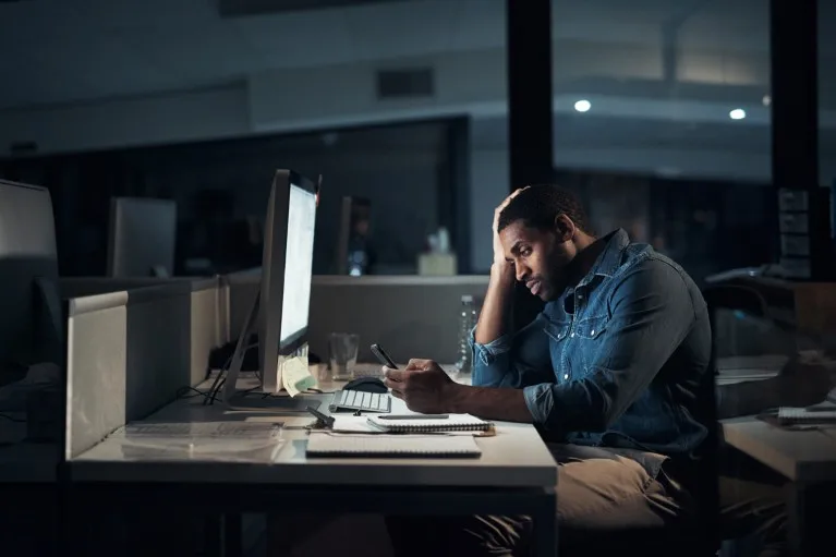 A man working at a computer in an office at night and looking at a smartphone