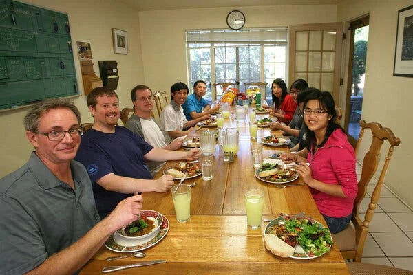 A dozen figures sit eating along a narrow wooden dining table. A large window in the back looks out onto treetops.