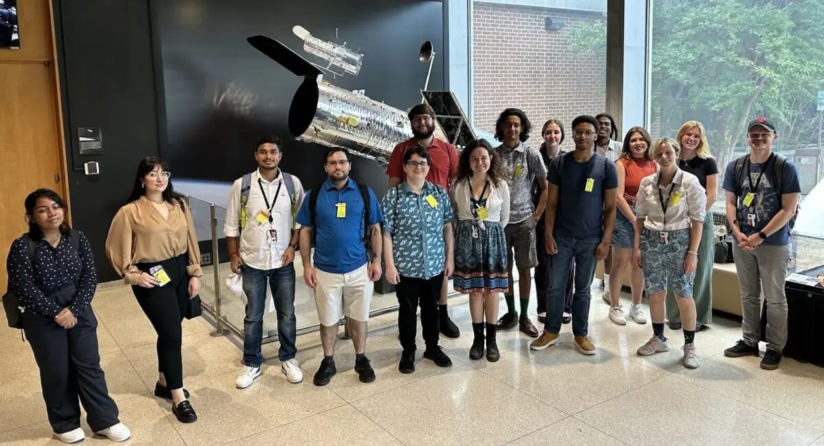 group photo in front a silver model of a telescope, about the size of a car