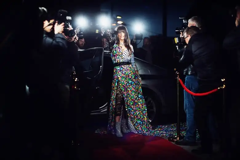 Image of a woman with long brown hair in a multi-partnered dress getting out of a limousine as photographers take images.