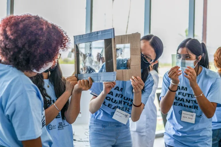 Female students in matching blue t-shirts look through cardboard frames with clear lenses as part of an experiment at a summer camp for quantum computing