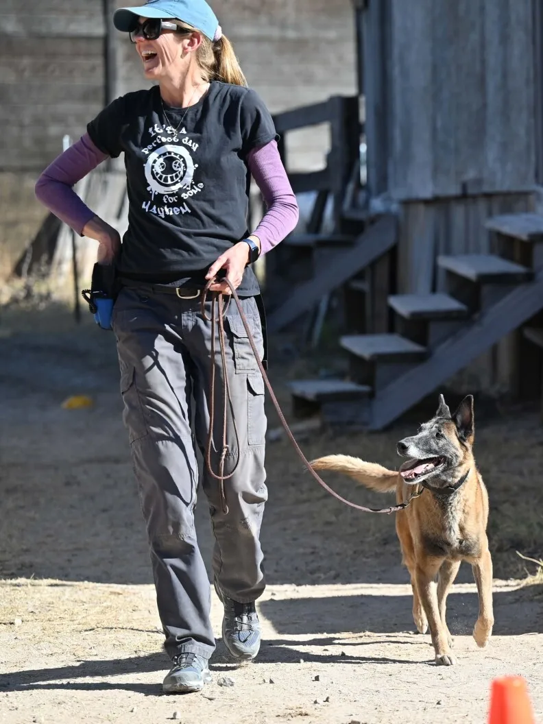 A woman wearing sunglasses and hat laughing at something to her left. A dog is standing next to her on leash, looking at her.