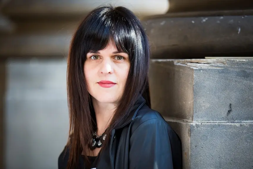 A white woman with dark brown hair and a fringe, standing against a bluestone wall and gazing at the camera