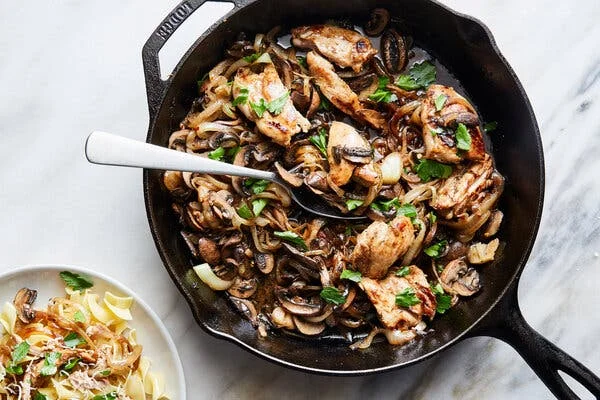 An overhead image of a cast-iron skillet filled with mushrooms, onions and chicken. A spoon scoops a bit of the mixture, and a single serving over egg noodles sits off to the side.