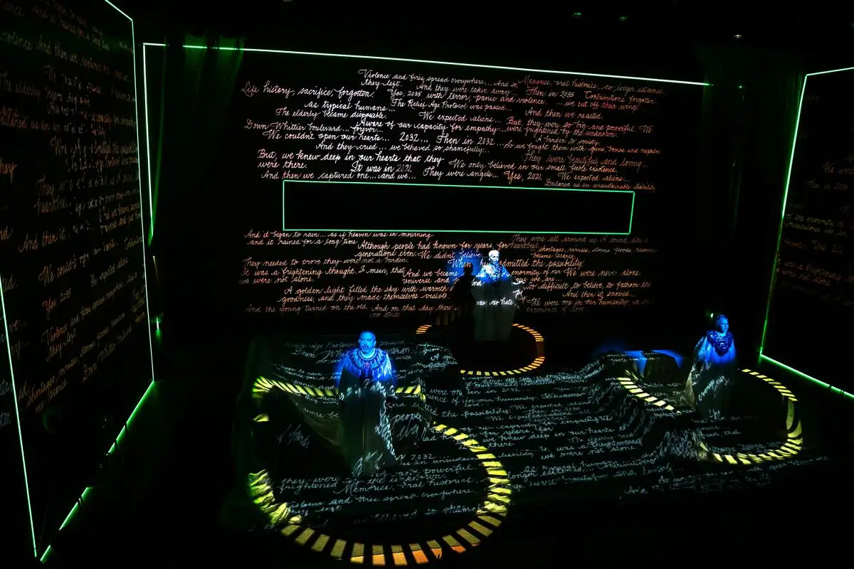 Three people sit on a darkened stage in circles of light, surrounded by screens of computer commands