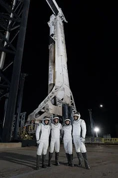 Four astronauts wearing white suits and helmets stand in front of a rocket on a launchpad.