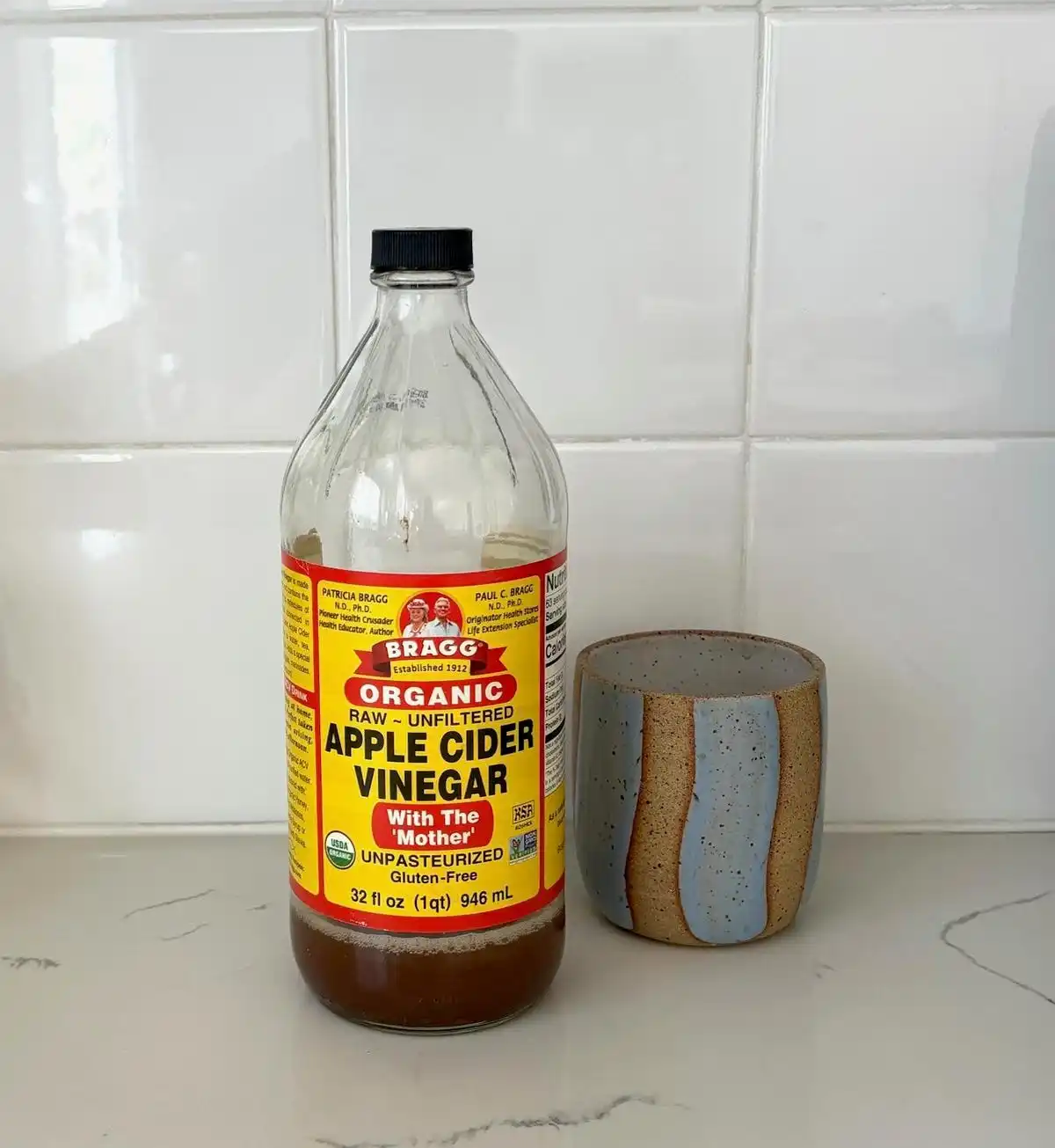 A bottle of Bragg organic apple cider vinegar next to a wavy blue and brown ceramic cup on a marble kitchen counter.