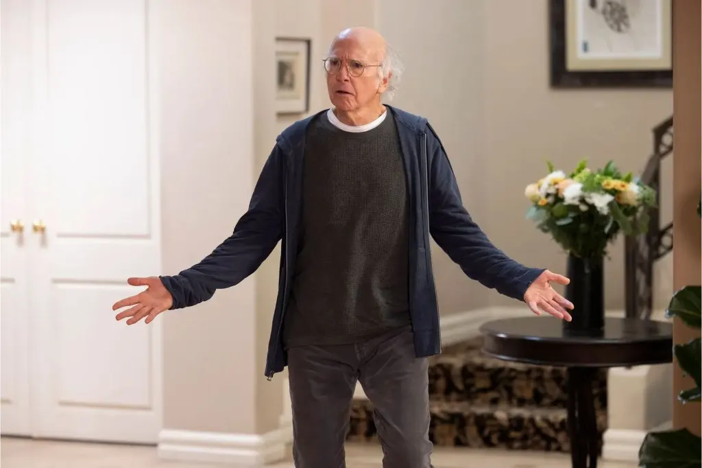 Larry David with his arms spread out near a curb