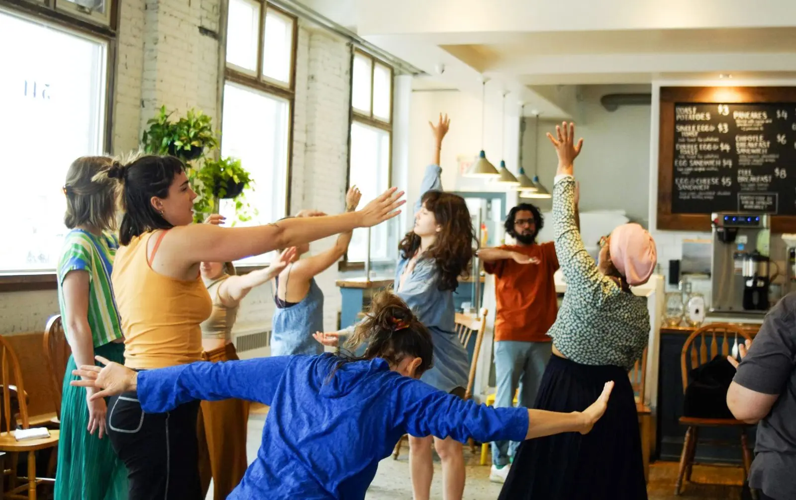 A group of people stand in a room with white brick walls and a coffee counter in the background, moving together with arms in the air and people in various positions invoking a sense of improvisation.