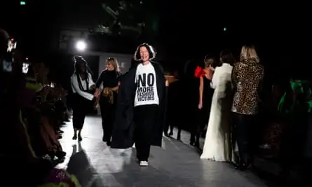 Woman on catwalk wearing black trousers and black coat with a white T-shirt with slogan ‘no more fashion victims’