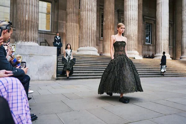 A model walking an outdoor runway in a black dress with a sheer bodice and a full skirt.