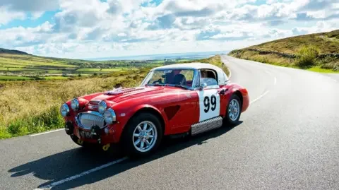 IOM FESTIVAL OF MOTORING A red classic car with the number 99 painted on the side driving along a country road. 