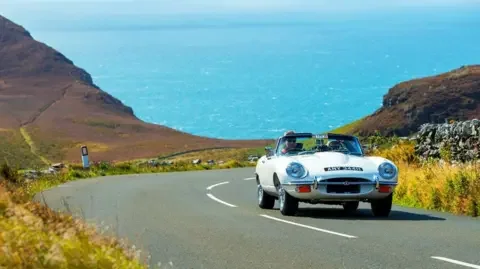 IOM FESTIVAL OF MOTORING A white classic convertible driving up a hill with coastline cliffs and the turquoise sea in the background.