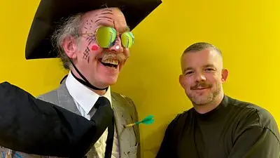 Image featuring two people. Russell Tovey smiles to camera, beside a man wearing sunglasses, a large hat, a novelty tie, and a paint-splattered suit. They have drawings on their face, and a fake dart protruding from their chest. 