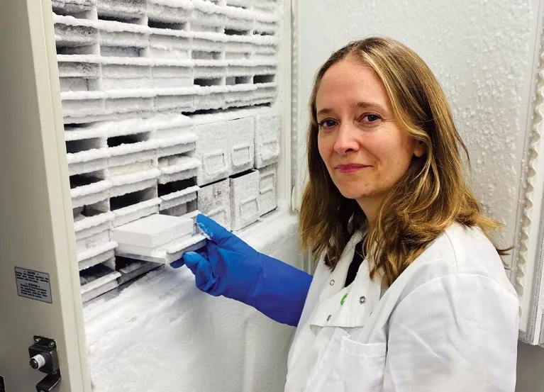 Joanne Durgan, wearing a white lab coat and blue gloves, looking to camera. She is extracting a sample from an ultra low temperature freezer.