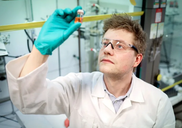 Thomas Freese in white lab coat and safety goggles is holding up a vial containing orange liquid in his green-gloved hand.