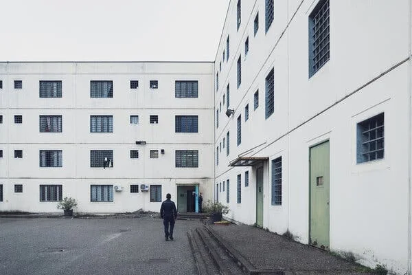 A man walking outdoors in a paved lot at the intersection of two exterior walls of a cream-colored building with bars on the windows. 