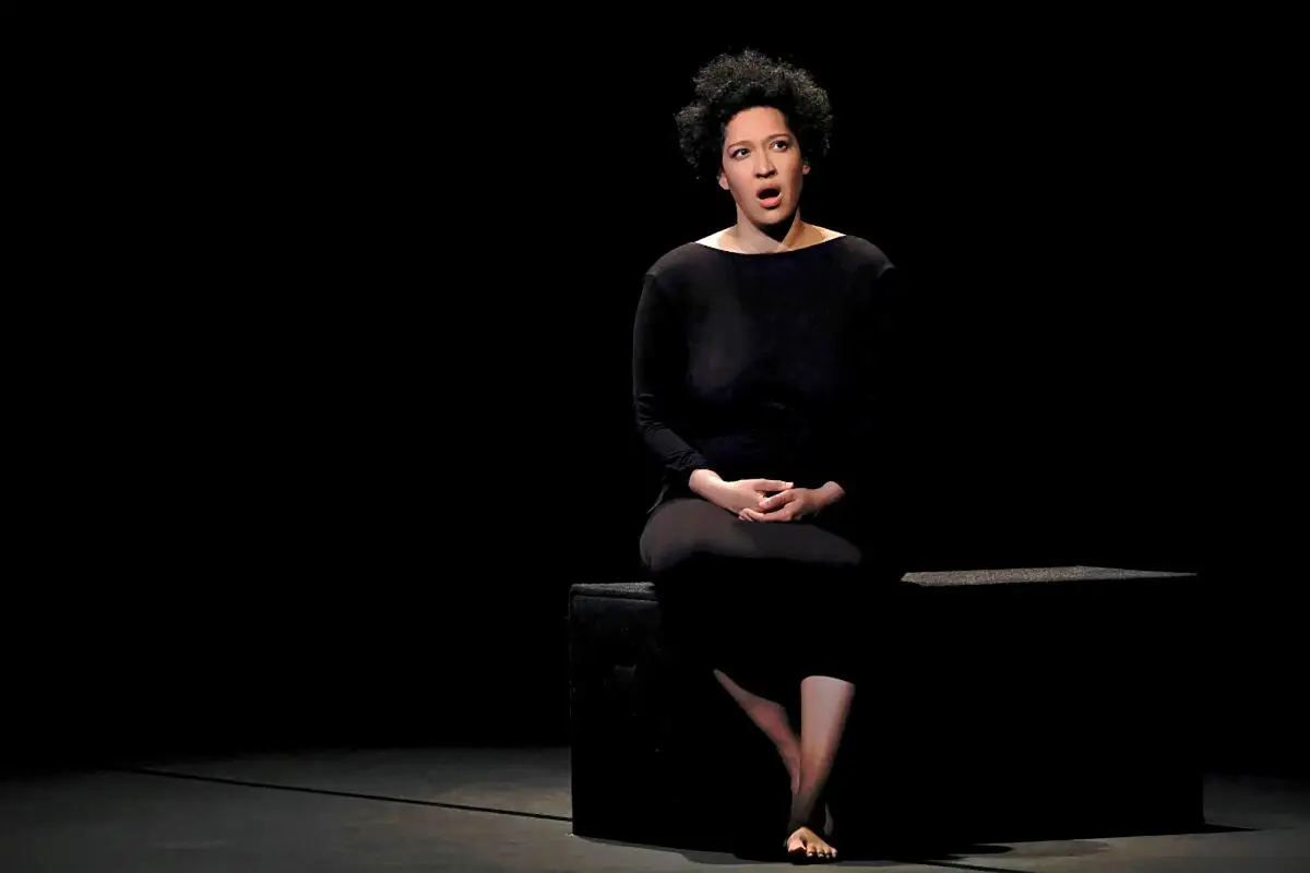 A woman singing while sitting on a black box on a darkened stage.