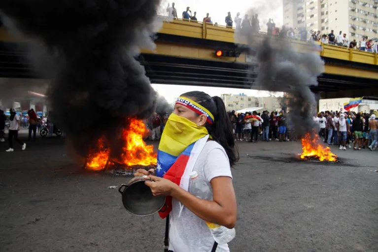 Fires burn behind a woman wearing a Venezuelan flag around her face, banging a pot during a protest in Puerto La Cruz