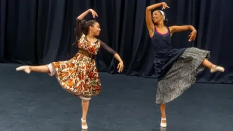 Oxygen Arts CIC Two black women performing ballet on stage. They are both wearing long dresses and looking towards each other. They are posed forming shapes with their arms and kicking one leg out while standing on their toes.