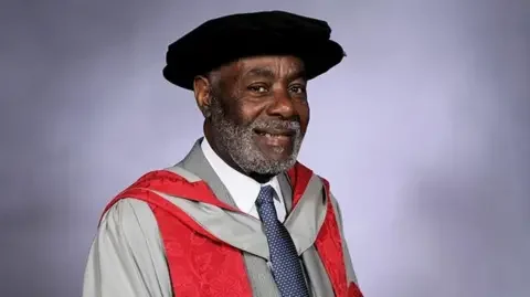 UWE Bristol Guy Bailey's graduation photo. He is wearing grey robes with a red lapel and a black PhD cap. He is standing against a blurry purple-grey background. 