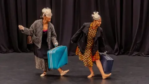 Oxygen Arts CIC Two black women dancing on stage in front of a curtain. One is dressed in a colourful African print and the other is wearing a long dress and padded jacket. They are both wearing beige fascinators in their hair and carrying suitcases.