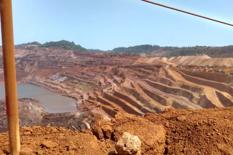 Image shows a deep mining pit surrounded by layers of dug-up soil and trees in the background