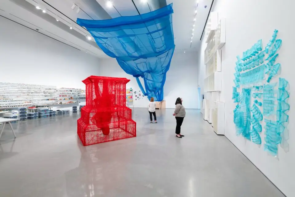 a massive blue fabric suspends from the ceiling while a red string covered structure stands in the gallery. a smaller light blue work is on the wall to the right