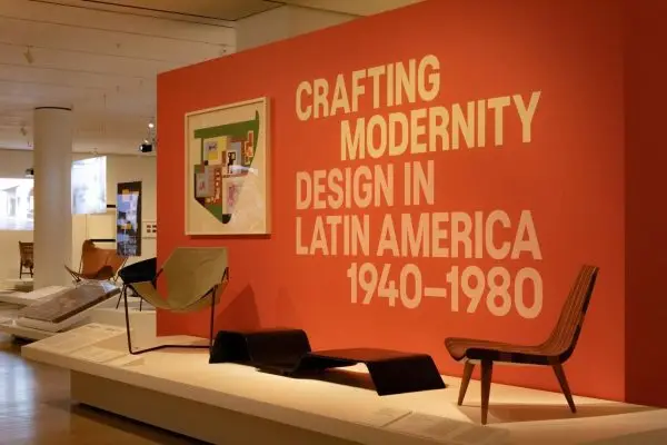 An exhibit featuring modern furniture on a stage with a large red panel that says “CRAFTING MODERNITY DESIGN IN LATIN AMERICA 1940-1980.”